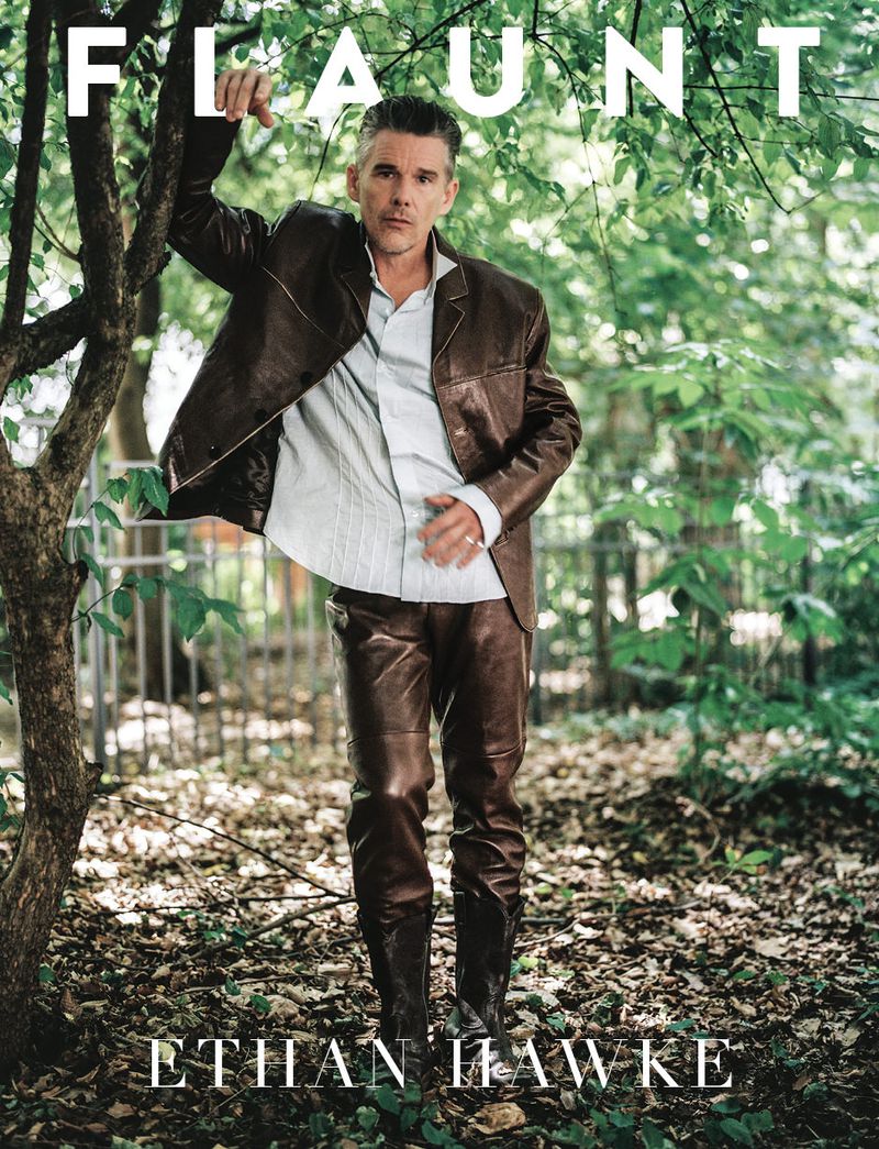 Ethan Hawke poses in a brown leather jacket and pants against a leafy outdoor background for a magazine cover, creating a stylish portrait.