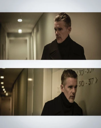 Portrait of a silver-haired man in a black turtleneck and coat standing in a hallway with neutral-colored walls and ceiling lights.