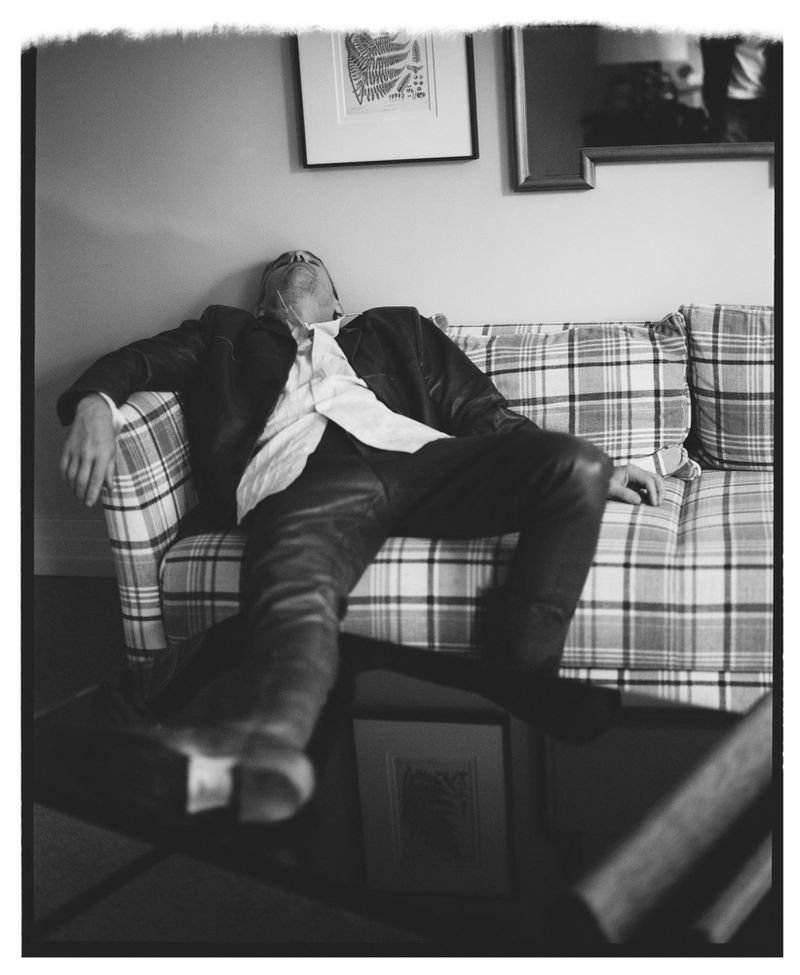 Black and white photo of Ethan Hawke reclining back on a plaid sofa in a leather jacket and button-down shirt.
