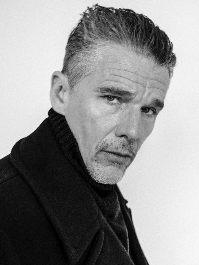 Black-and-white portrait of a man with salt and pepper hair wearing a turtleneck and dark coat, gazing into the camera.