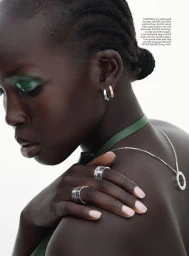 Model Ajok Madel in profile wearing silver and gold Cartier rings, earrings, and green eyeshadow against a white background.