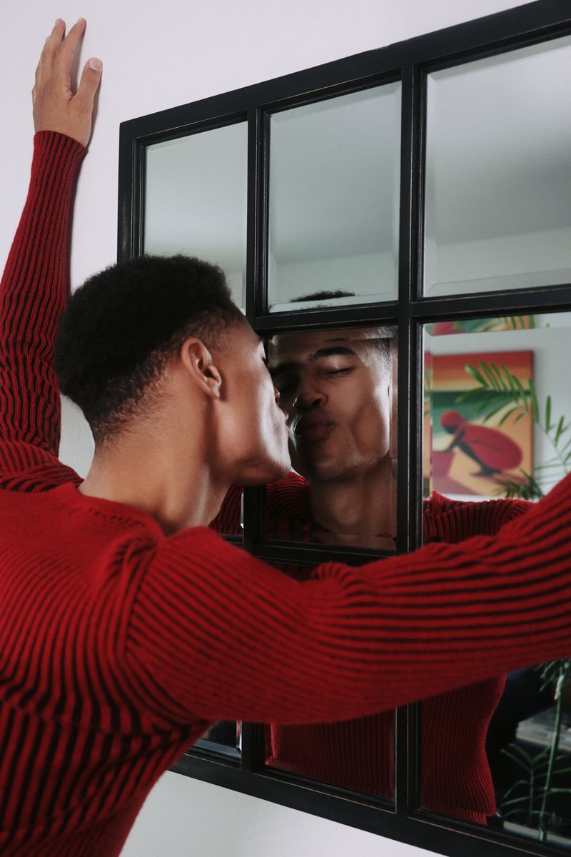 An adult Black male in a red ribbed sweater kisses his reflection in a windowpane-style mirror, indoors with a colorful painting in the background.