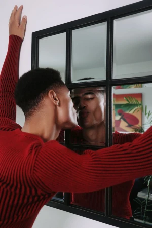 An adult Black male in a red ribbed sweater kisses his reflection in a windowpane-style mirror, indoors with a colorful painting in the background.