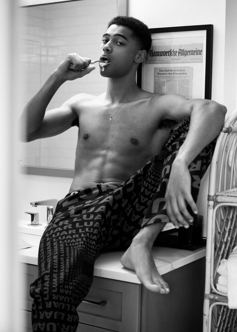 Young adult male brushes teeth while sitting on bathroom sink wearing patterned pants, newspaper in background, black and white studio shot.
