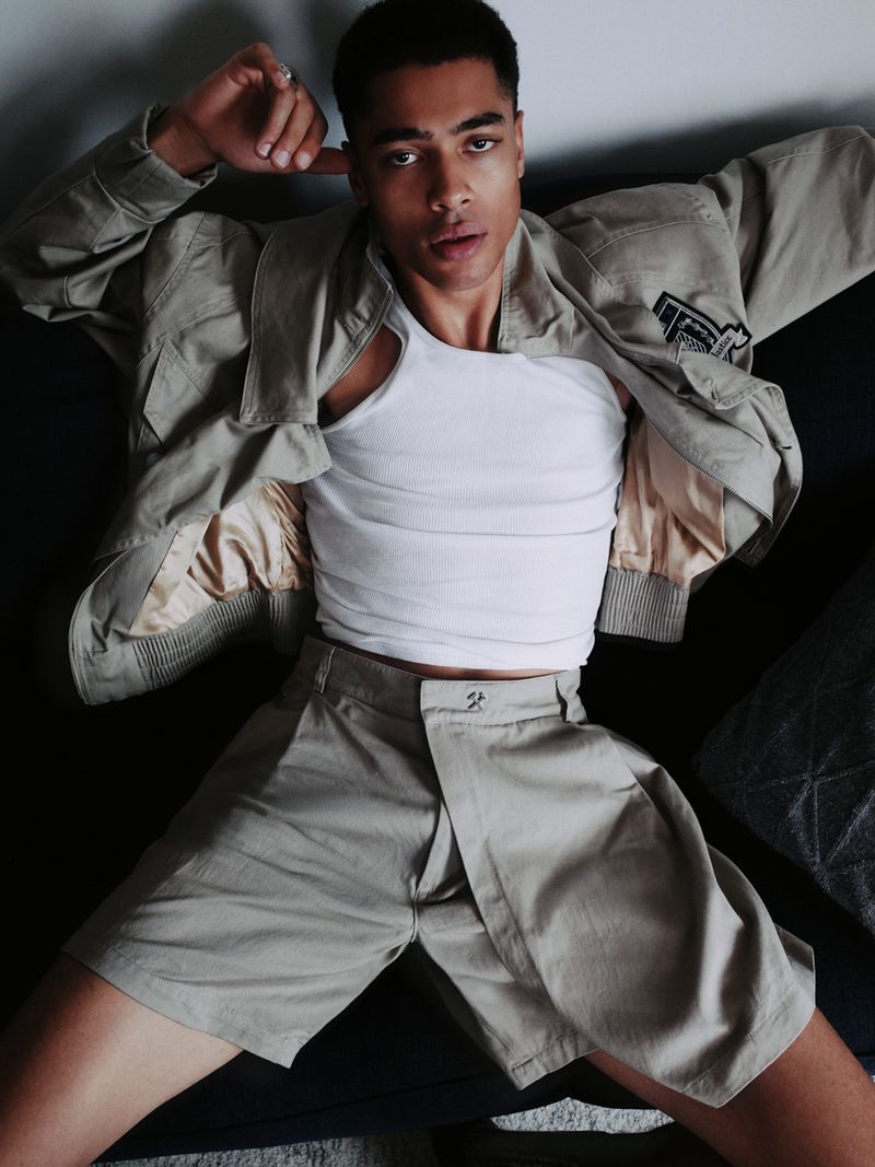Portrait of a Black male model wearing a white tank top, khaki jacket, and shorts, lying down indoors looking directly at the camera.