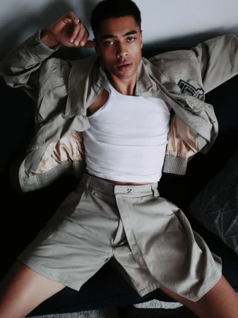 Portrait of a Black male model wearing a white tank top, khaki jacket, and shorts, lying down indoors looking directly at the camera.