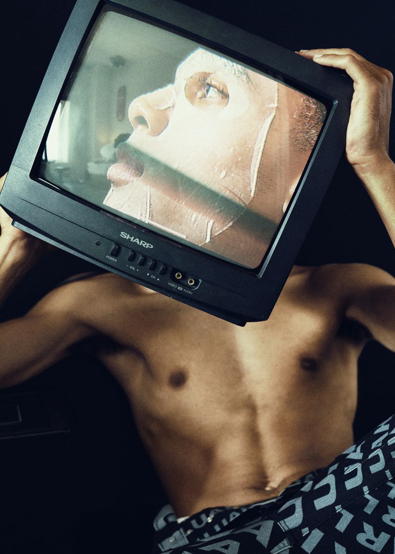 Shirtless person with facial mask and printed pants holding a small television in front of their face against a black background.