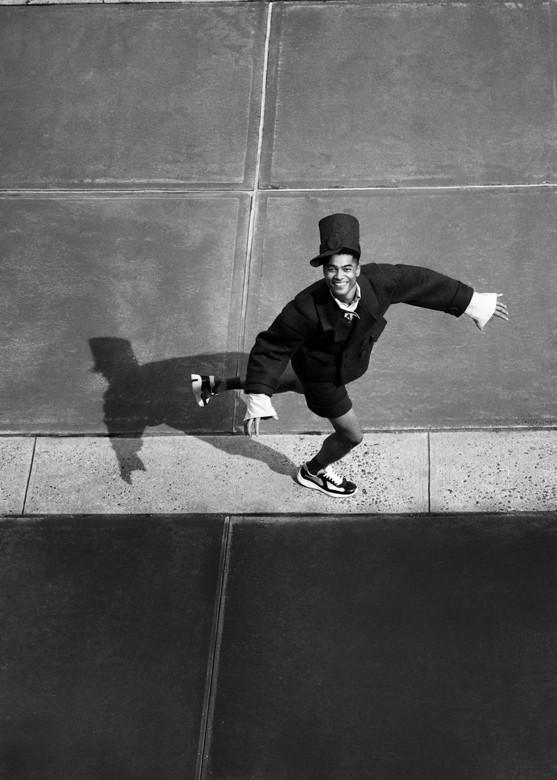 High-angle black and white shot of a man wearing a top hat, dark jacket, and shorts leaping across paved concrete slabs.