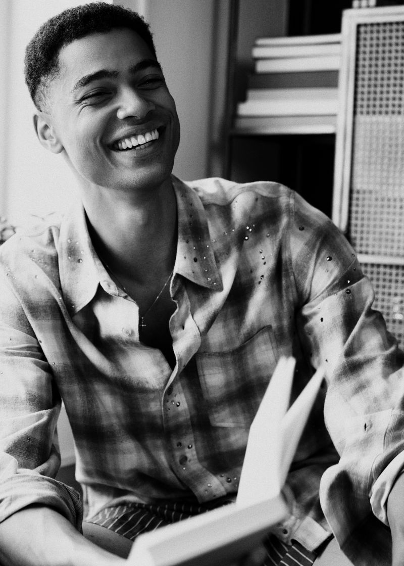 Désiré Mia laughing and holding an open book while wearing a plaid button-up shirt in a black and white editorial portrait.