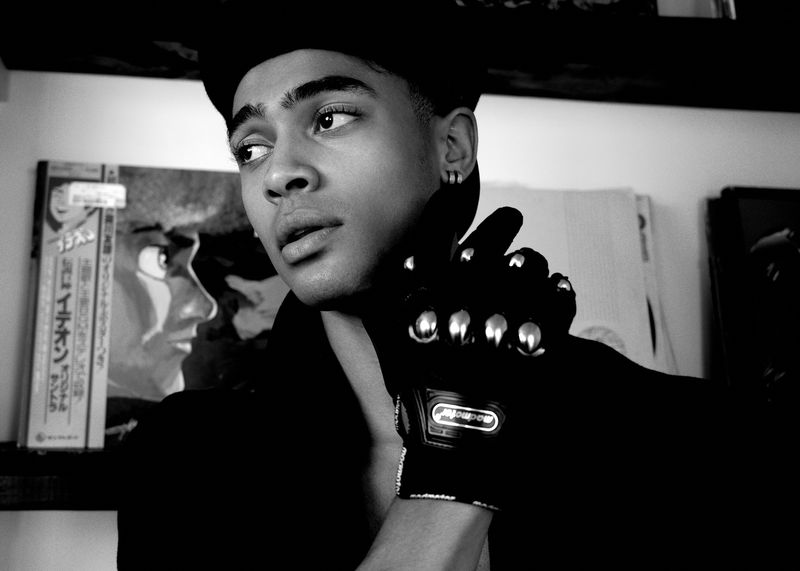 Monochrome portrait of a young adult looking left, wearing a cap and black clothing, with a studded black glove visible.
