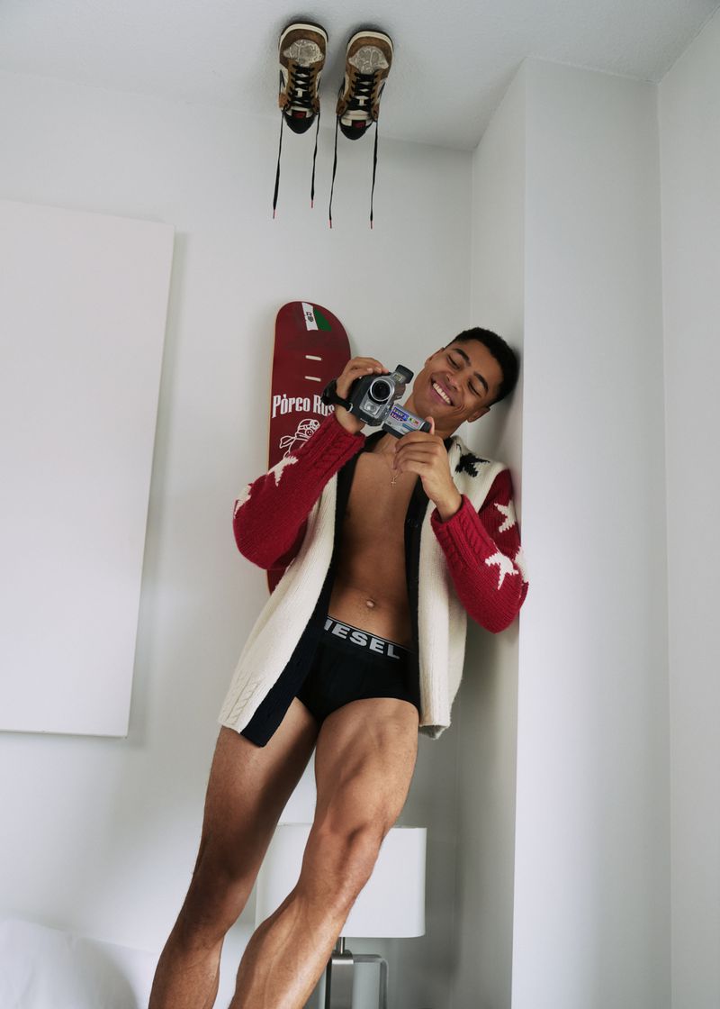 Smiling adult man with vintage camera indoors, wearing a red and white sweater, posing in underwear. Pairs of shoes on ceiling.