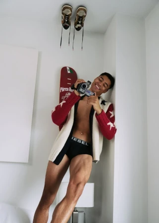 Smiling adult man with vintage camera indoors, wearing a red and white sweater, posing in underwear. Pairs of shoes on ceiling.