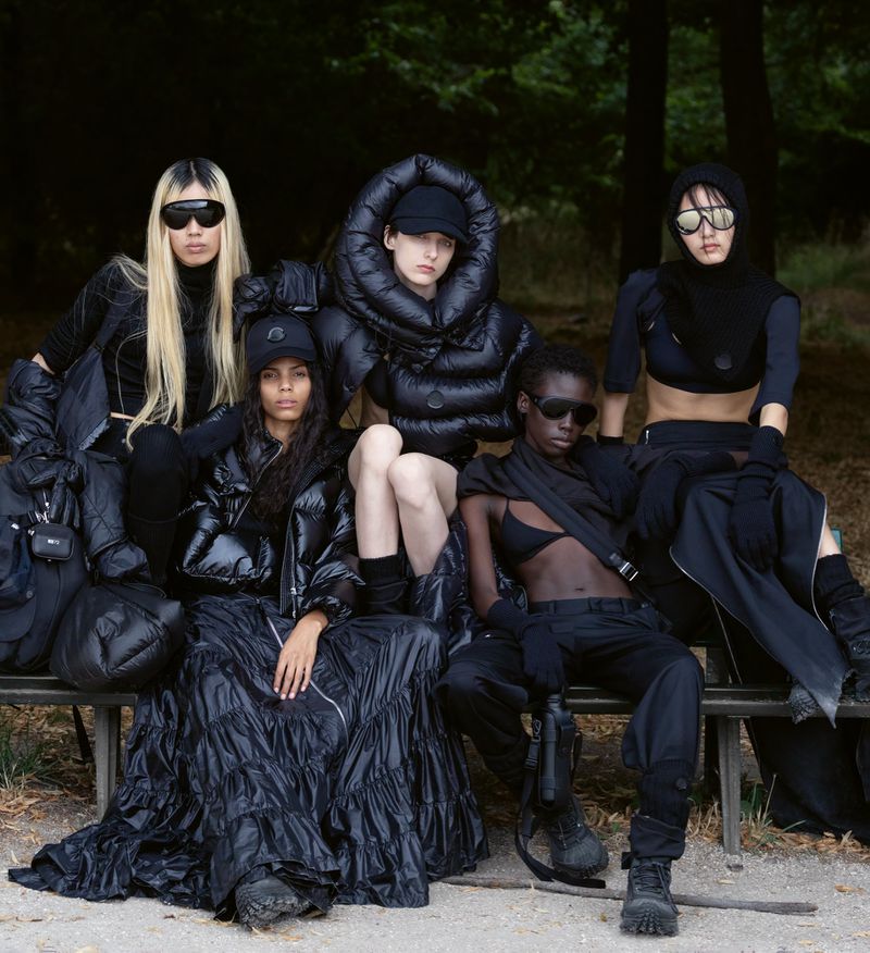 Five fashion models in all-black attire sit on a bench in an outdoor setting, showcasing a cohesive fashion collection with puffer jackets.