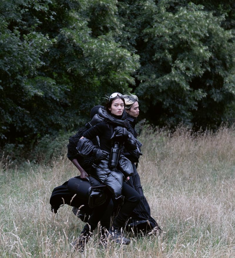 Two women in matching all-black outfits and goggles walking through tall grassy field with dark green trees in background on overcast day.