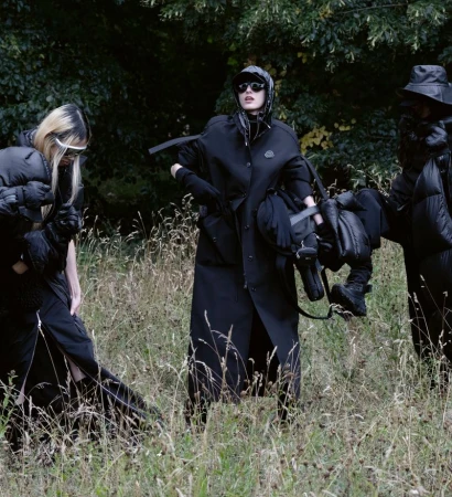 Three models in dark apparel pose in a field of tall grass, with trees and foliage in the background, creating a dark, stylish outdoor shot.