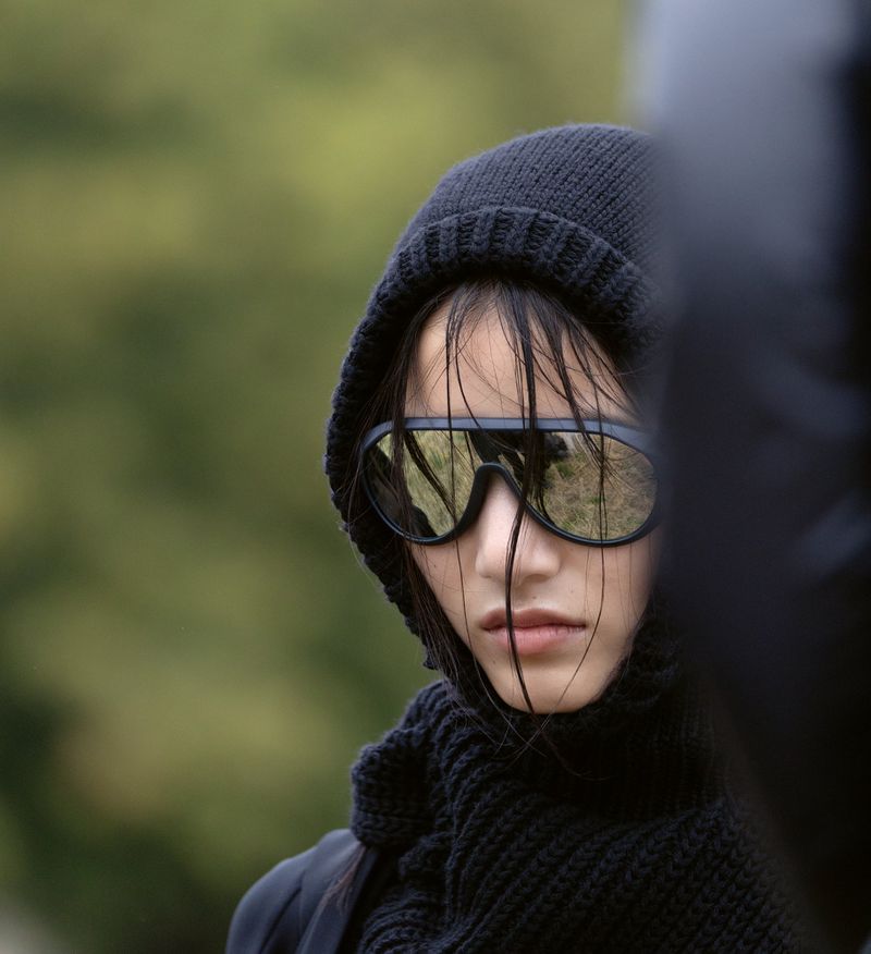 Portrait of an Asian adult female wearing dark sunglasses, a knitted beanie, and a scarf against a blurred green background.