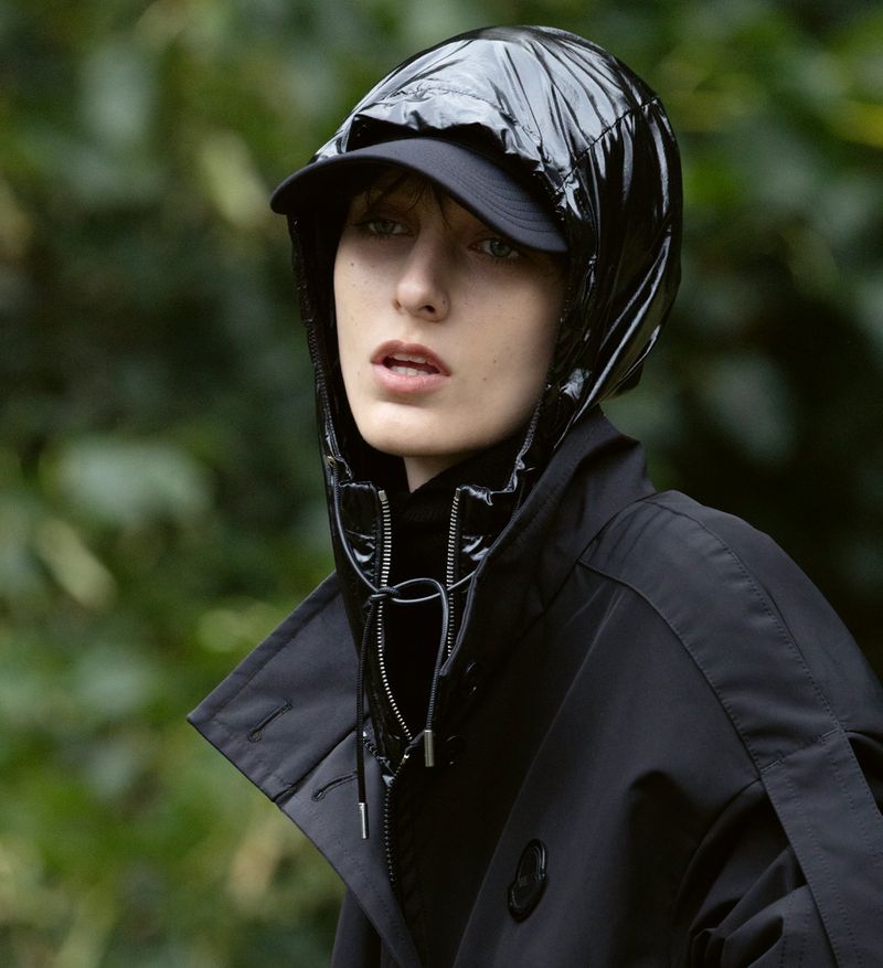 Woman with light skin, wearing a black coat and a shiny black hooded hat poses in front of a blurred green background outdoors.