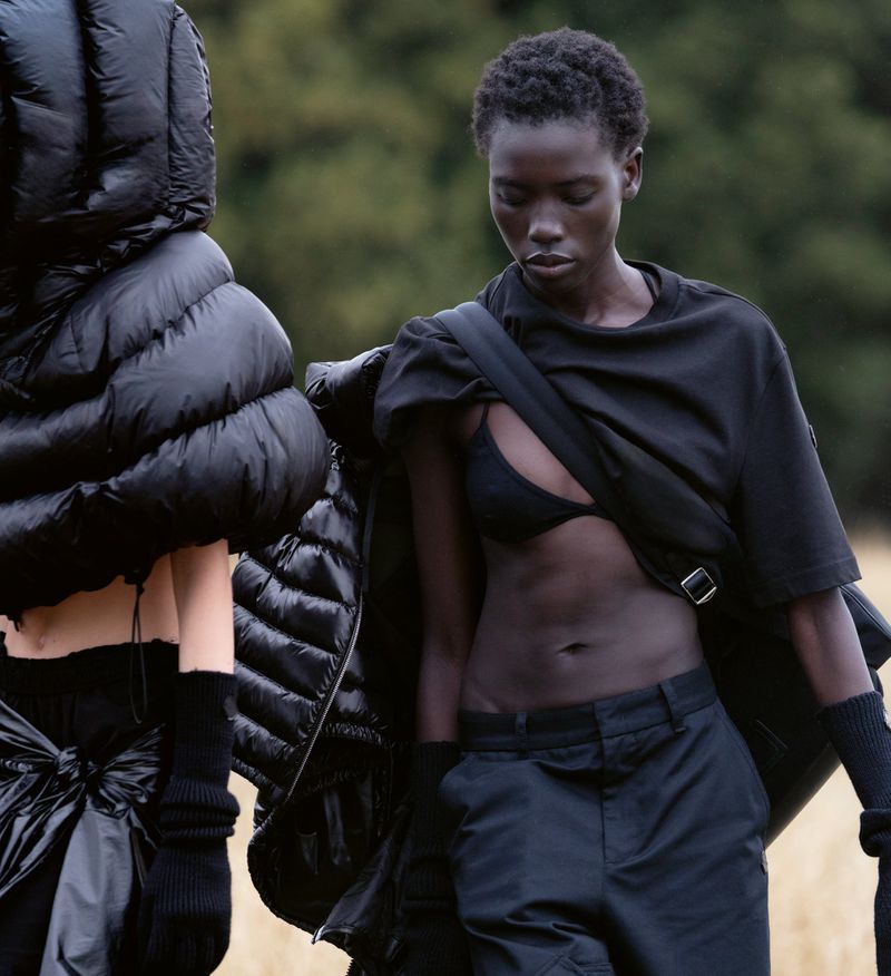 Two models wearing all-black fashion pose outdoors in a field. One wears an unzipped puffy coat, the other a t-shirt and bra top with cargo pants.
