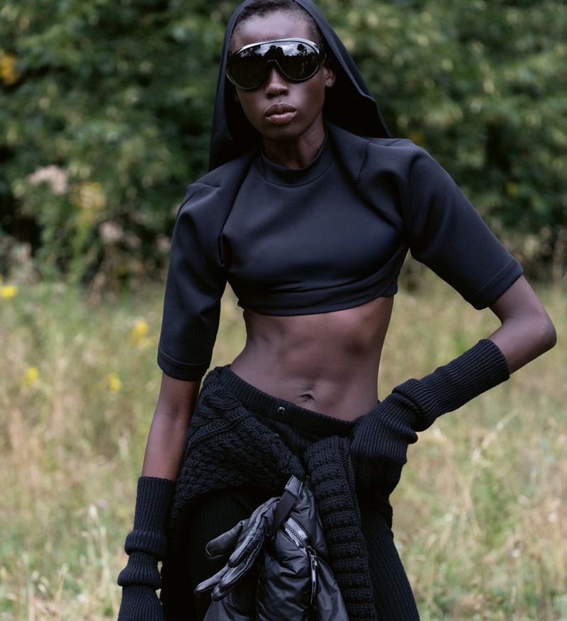 A woman with dark skin and wearing a black hooded outfit, sunglasses, and knitwear poses outdoors amid blurred green foliage.