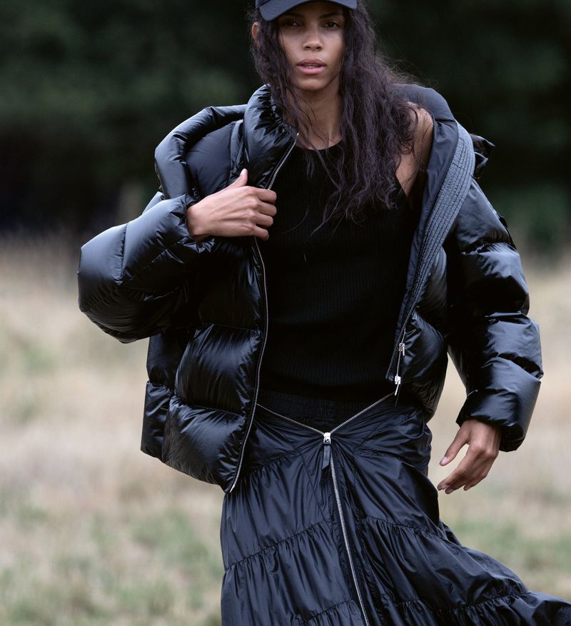 An adult female poses in an outdoor setting wearing a black baseball cap, black top, black puffer jacket, and a black tiered skirt.