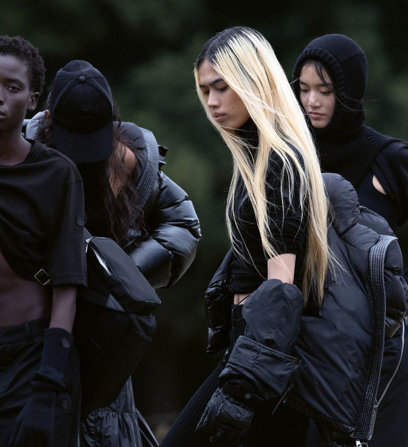 A group of young adult fashion models in dark-colored contemporary clothing stand together outdoors during daytime, posing in neutral expressions.