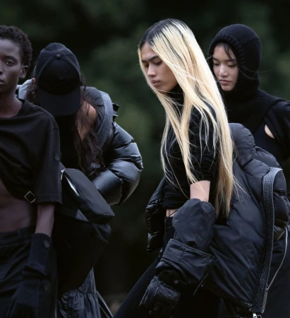 A group of young adult fashion models in dark-colored contemporary clothing stand together outdoors during daytime, posing in neutral expressions.