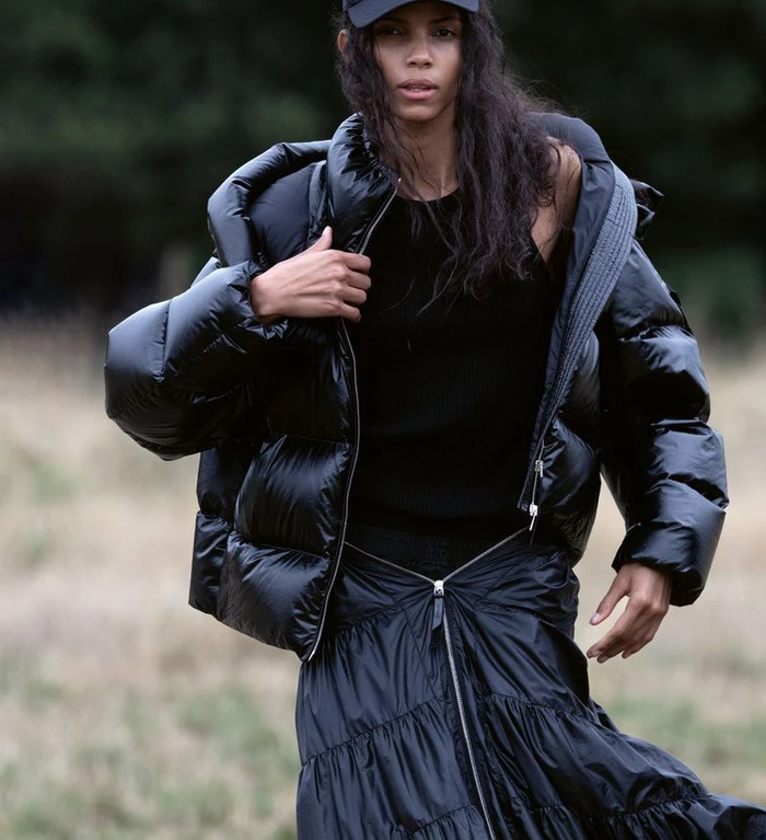 Model wearing a glossy black puffer jacket and dark cap standing against a blurred natural outdoor background