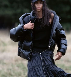 Model wearing a glossy black puffer jacket and dark cap standing against a blurred natural outdoor background