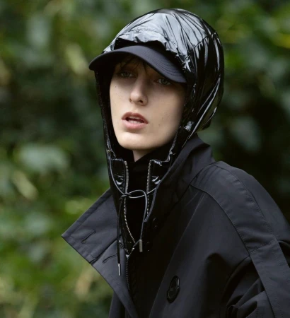 Model wearing a glossy black hooded jacket over a black cap against a soft focus green foliage background.