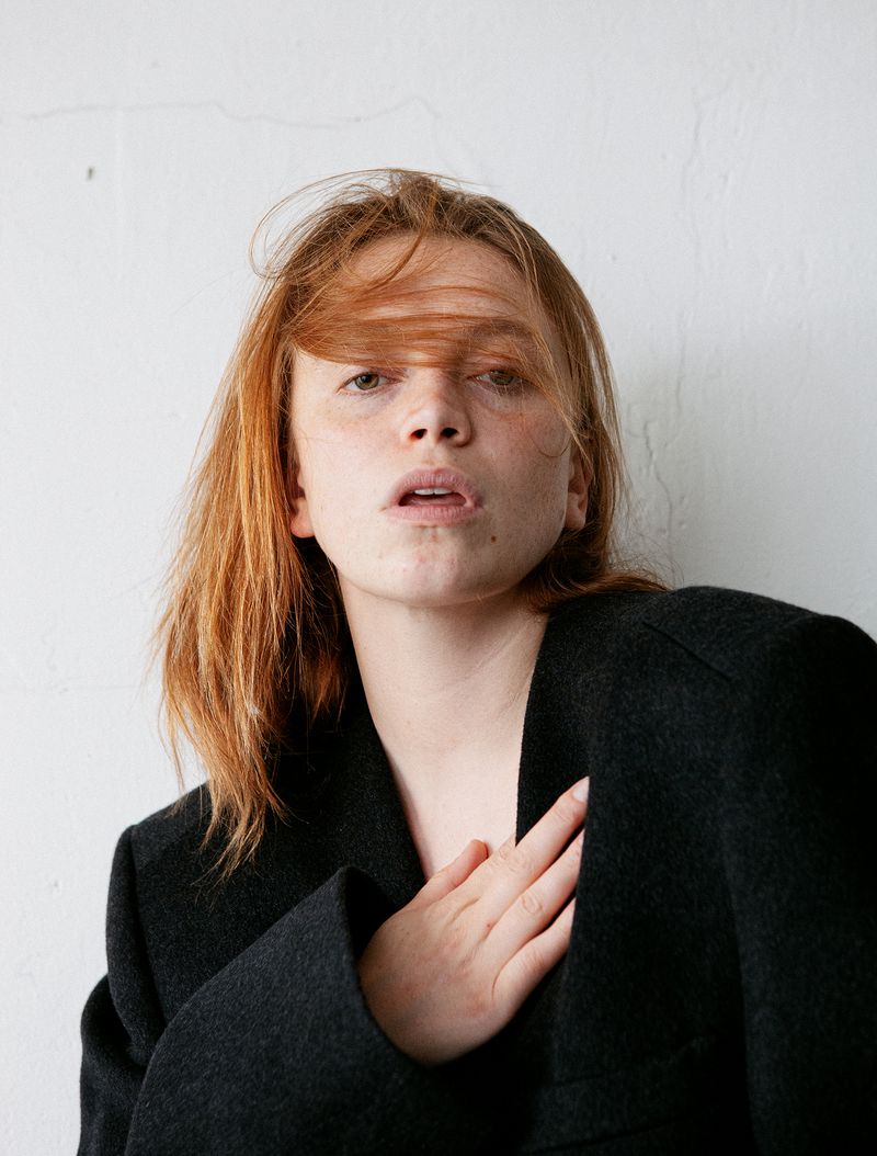 Close-up portrait of a red-haired female model with fair skin wearing a dark coat, looking at the camera against a white background.