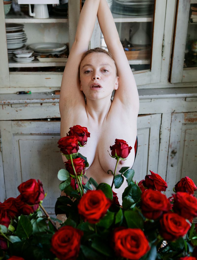 Artistic portrait of a fair-skinned woman with arms raised, partly obscured by bouquet of red roses, in front of vintage cupboard.