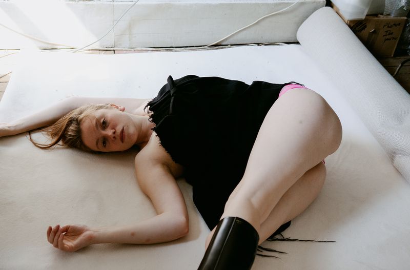 Red-haired woman poses in a black dress on a white backdrop, wearing shiny black boots, under bright, diffused lighting in a studio setting.