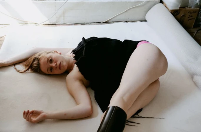 A female model lying down on a white studio paper sweep wearing a black textured fringed dress and black boots.