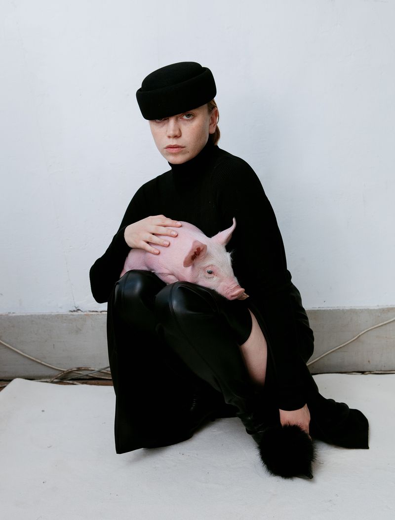 A woman in black turtleneck and black hat holds a pink piglet, seated on a white surface against a white wall, creating a minimalist aesthetic.