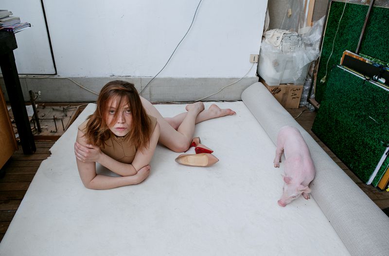 Young woman poses with a pink pig on a white carpet indoors. The room appears to be an artist's studio, with art supplies visible in the background.