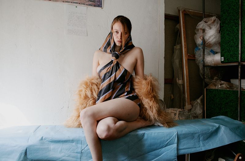Nude woman draped in a striped scarf and fur shawl sits on a blue-covered bed in a room with art supplies, creating an intimate and artistic image.