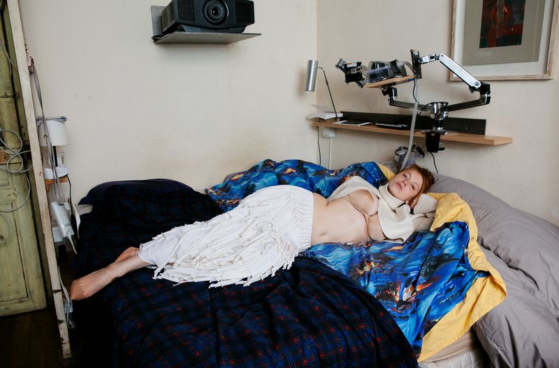 A woman reclines on a bed in an indoor setting, with a projector and camera equipment visible in the background. The light is soft.