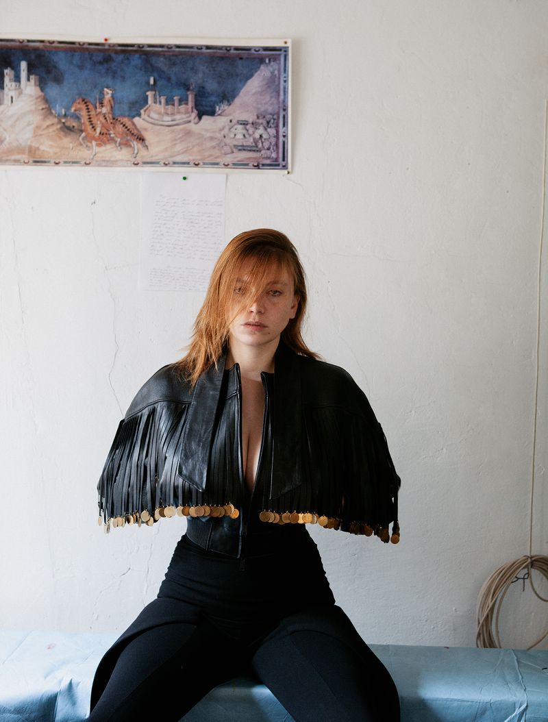 Portrait of a young woman with red hair wearing a black fringed jacket and pants, sitting indoors with a painting in the background.