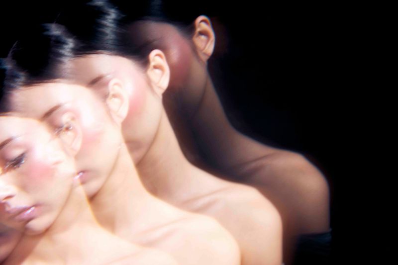 Dreamy portrait of a woman with blurred multiple exposures against a dark backdrop, exhibiting soft lighting and a serene aesthetic.