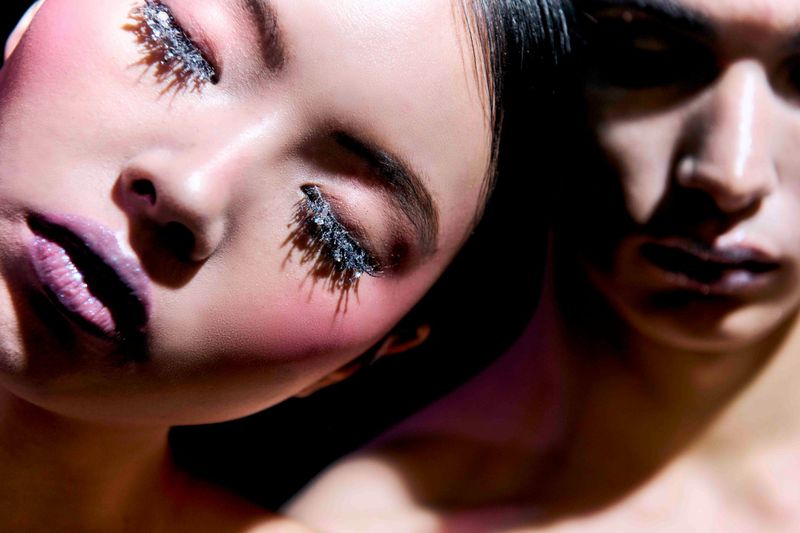 Close-up of two Asian adults with creative makeup, featuring embellished eyelashes and purple lipstick, under studio lighting in a dark setting.