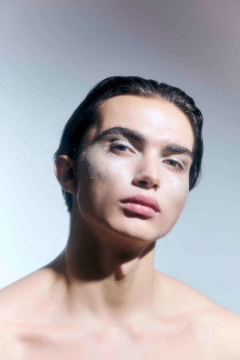 Portrait of a shirtless, young adult male with dark hair and light skin, wearing shimmering makeup, under soft, diffused lighting.
