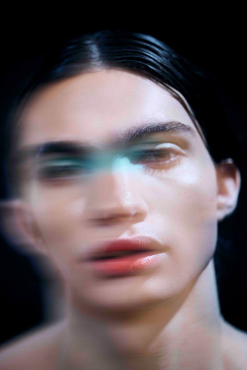 Close-up studio portrait of a person with slicked-back hair, red lipstick, and blurred facial features against a dark background.