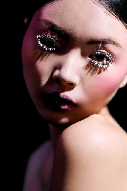 Close-up portrait of a woman with dramatic makeup, silver glitter eyelashes, dark lipstick, and dark hair against a black backdrop.