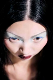 High-angle close-up portrait of a woman with dark hair, pale eyeshadow, blue eyelashes, and dark lipstick against a dark background.