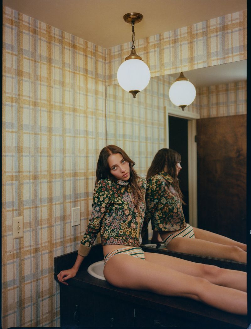 A young woman poses in a floral top and striped underwear, sitting on a sink counter in a retro-style bathroom with plaid wallpaper.