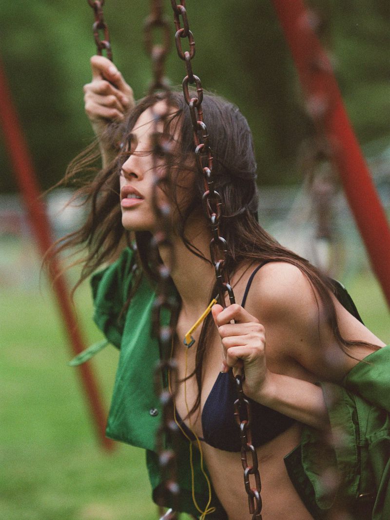 Woman in bikini and green jacket sits on a metal chain swing in an outdoor park setting, looking up with a neutral expression.