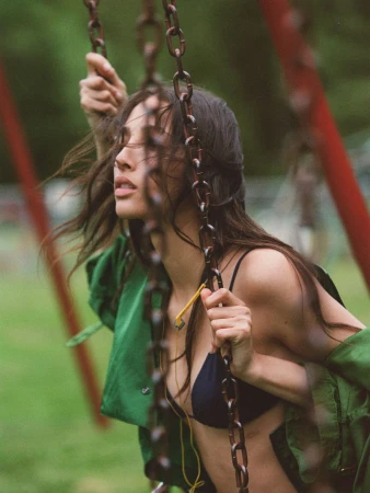 Woman in bikini and green jacket sits on a metal chain swing in an outdoor park setting, looking up with a neutral expression.