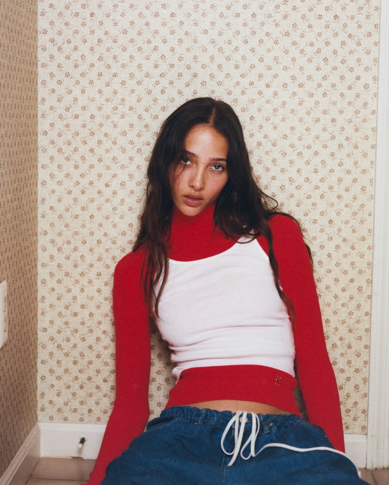 Young adult female with long brown hair poses indoors wearing a red long sleeve shirt, white tank top, and blue sweatpants.