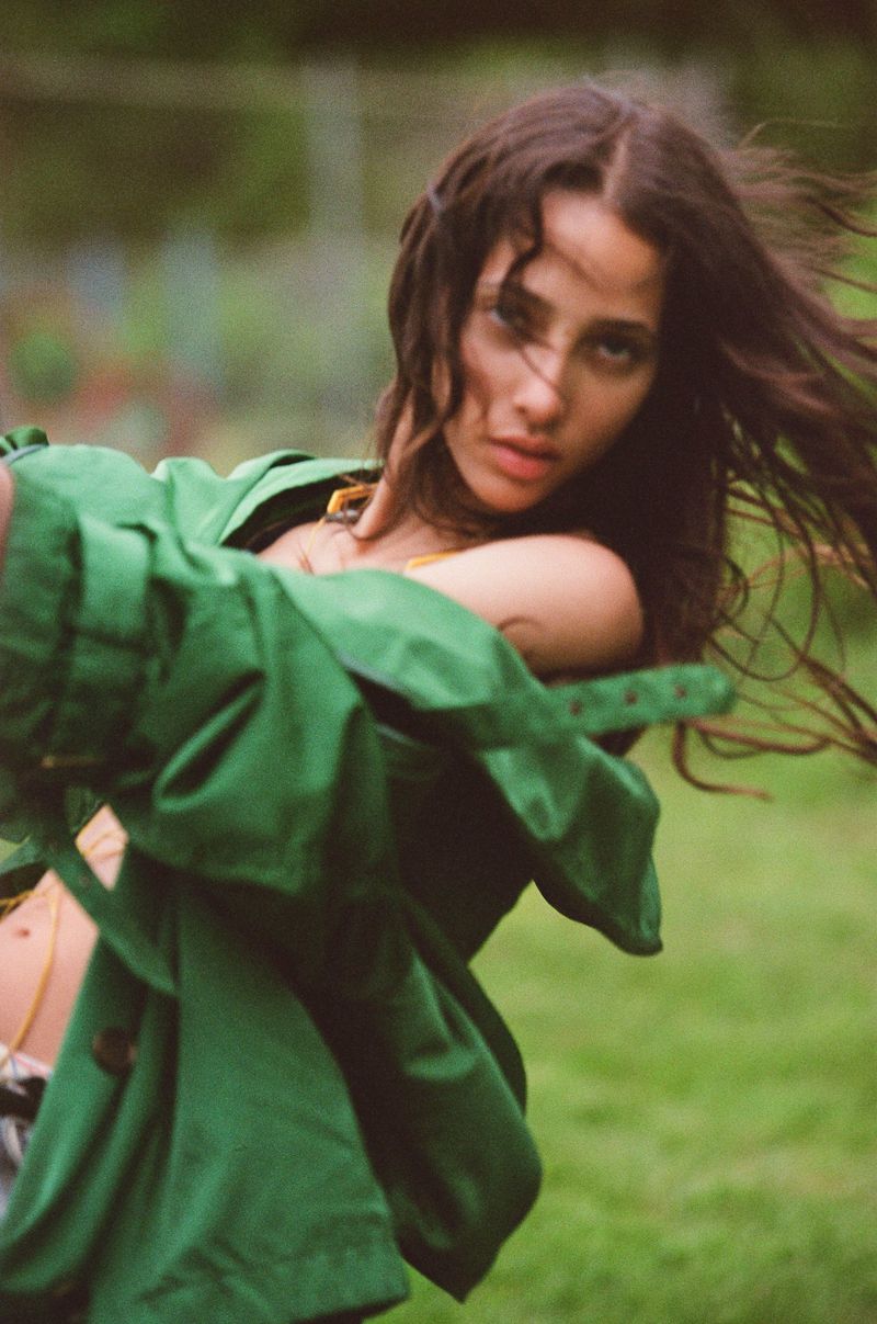 Young woman with long wavy hair wearing a green jacket in a natural outdoor setting with a soft, blurred background and shallow depth of field.
