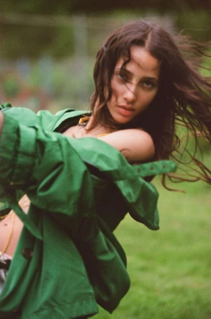 Young woman with long wavy hair wearing a green jacket in a natural outdoor setting with a soft, blurred background and shallow depth of field.